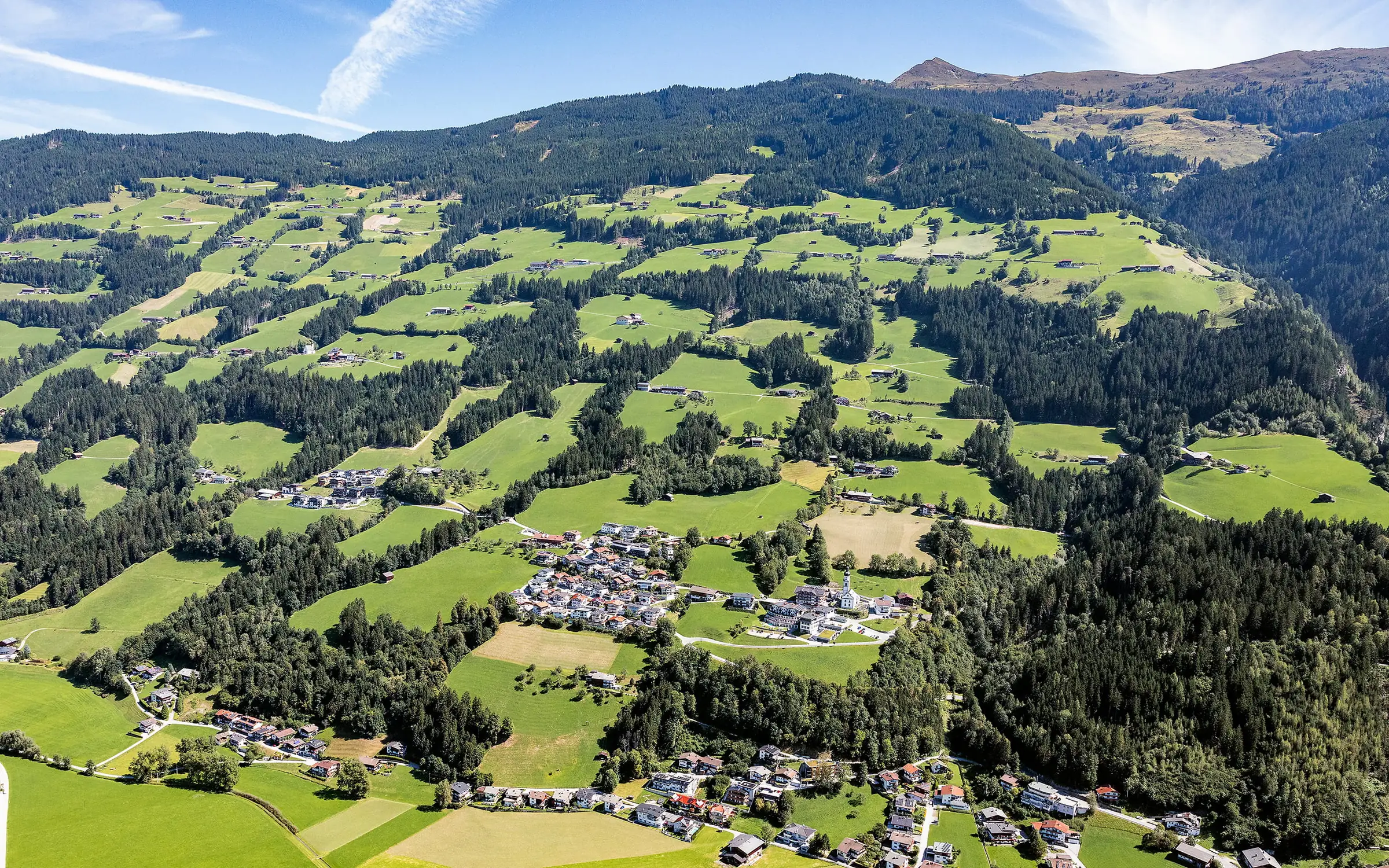 Luftbild von Hart im Zillertal: Streusiedlung mit Höfen und Wiesen am bewaldeten Hartberg, in der Mitte die Pfarrkirche.